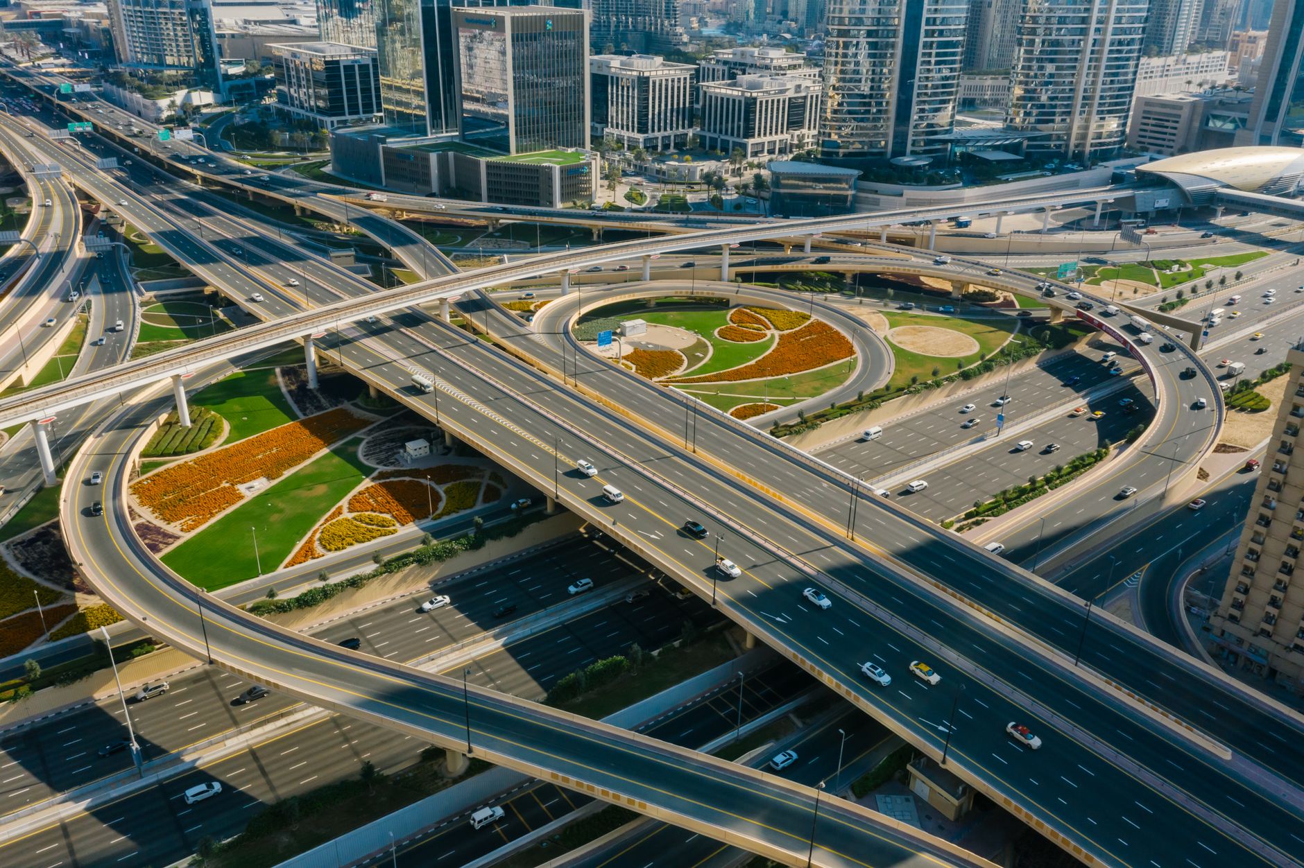 Aerial view of a multi-level motorway interchange in Dubai, with landscaped roundabout gardens, flowing traffic and high-rise towers beyond