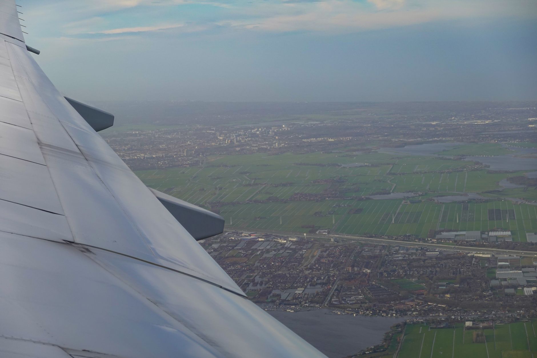 A plane's wing over green fields, a town and a lake, seen from a window seat.