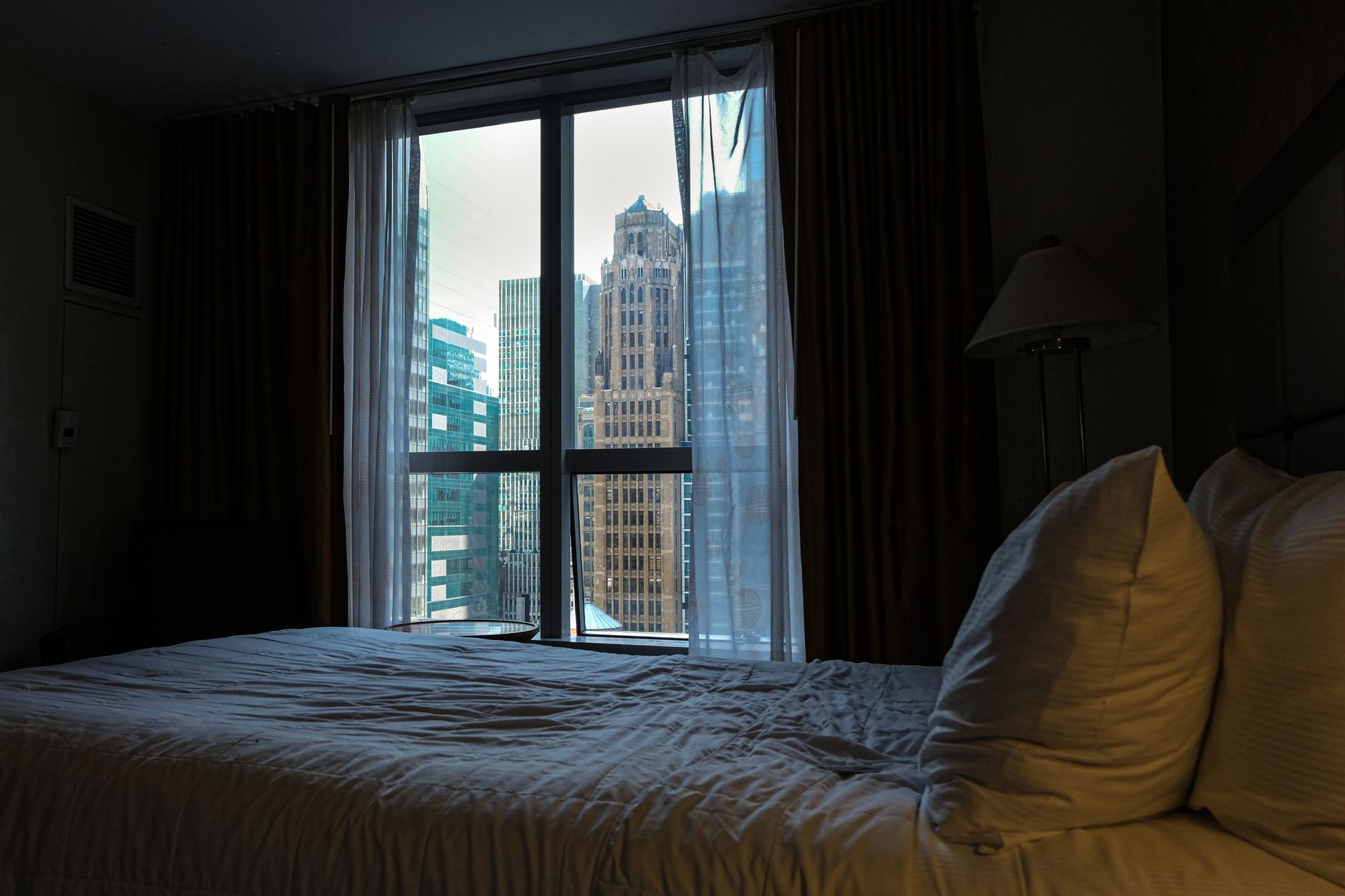 A dim hotel room with a bed in front of a tall window looking out on city towers by day.